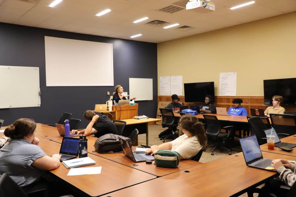 A teacher stands at a podium in a classroom, addressing students seated at tables with laptops. The atmosphere is engaged and focused.