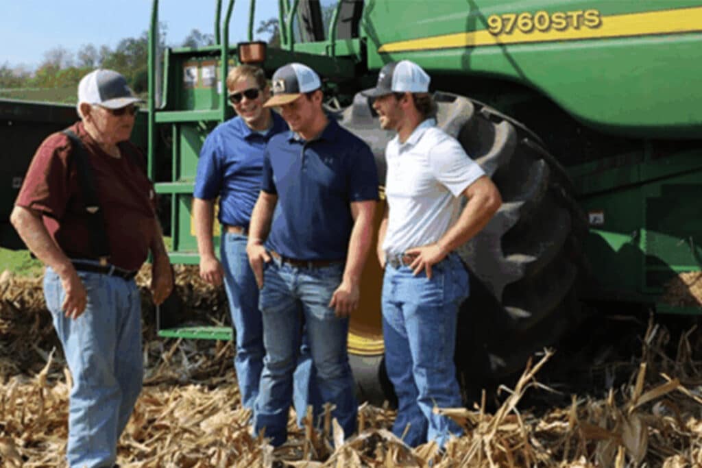 Group of men standing beside a large green combine harvester in a cornfield, discussing farm operations and agribusiness management.