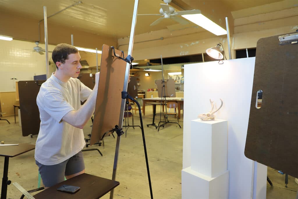 A young artist intently sketches on an easel in a well-lit art studio. A sculpted animal skull sits on a pedestal, illuminated by a small lamp.
