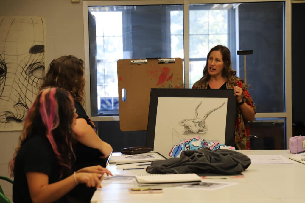 A woman presents a framed skill sketch to two seated students in an art classroom. The room has large windows and various drawings. The atmosphere is attentive.