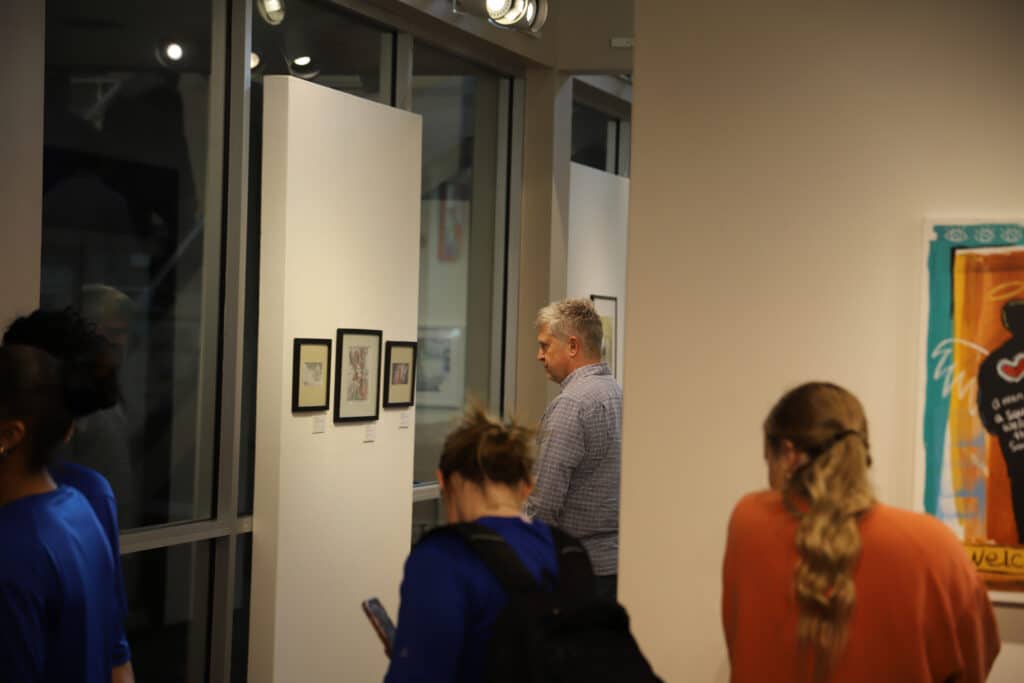 A group of people in a well-lit art gallery, absorbed in examining framed artworks on white walls. The scene conveys a quiet, contemplative atmosphere.