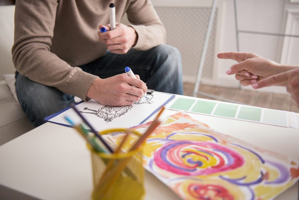 Close-up of two people engaged in drawing. One sketches a sheep while the other points, hinting collaboration. Bright, colorful artwork on the table. Creative mood.