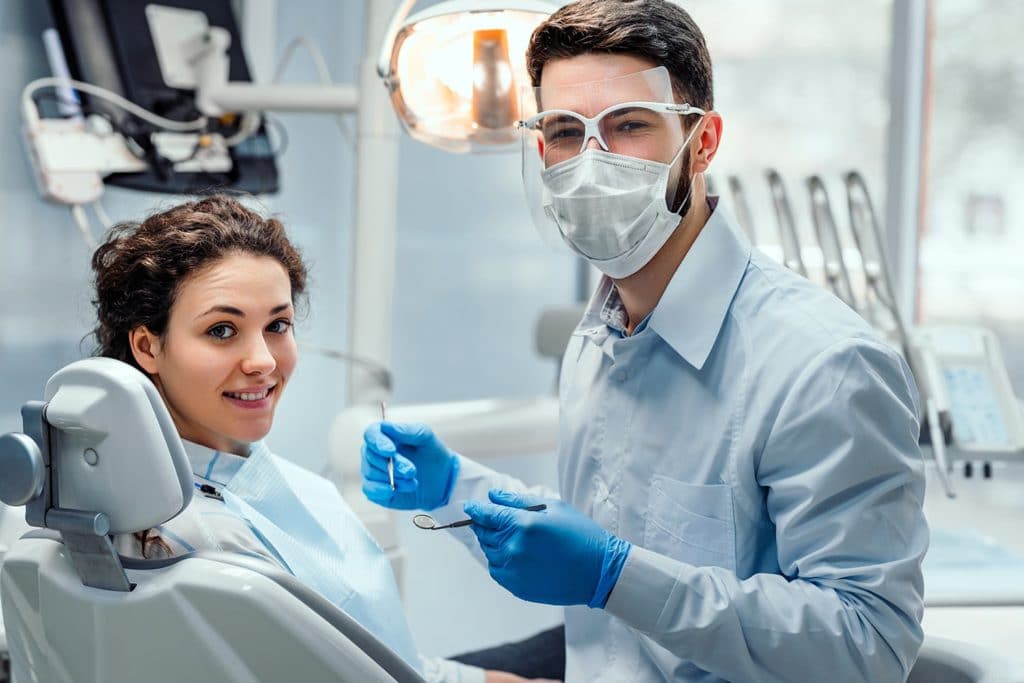 A dentist in protective gear, mask, and gloves holds dental tools, smiling alongside a relaxed patient in a dentist's chair, creating a calm setting.