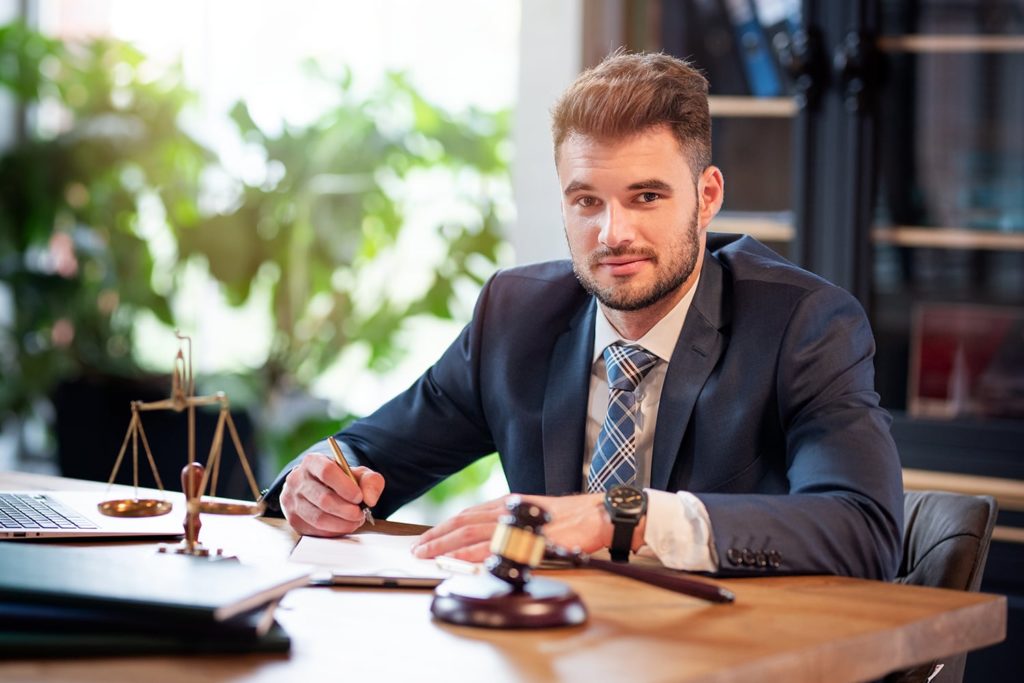 A man in a suit sits confidently at a desk with a gavel, legal scales, and a laptop. The setting is professional, with greenery in the background.