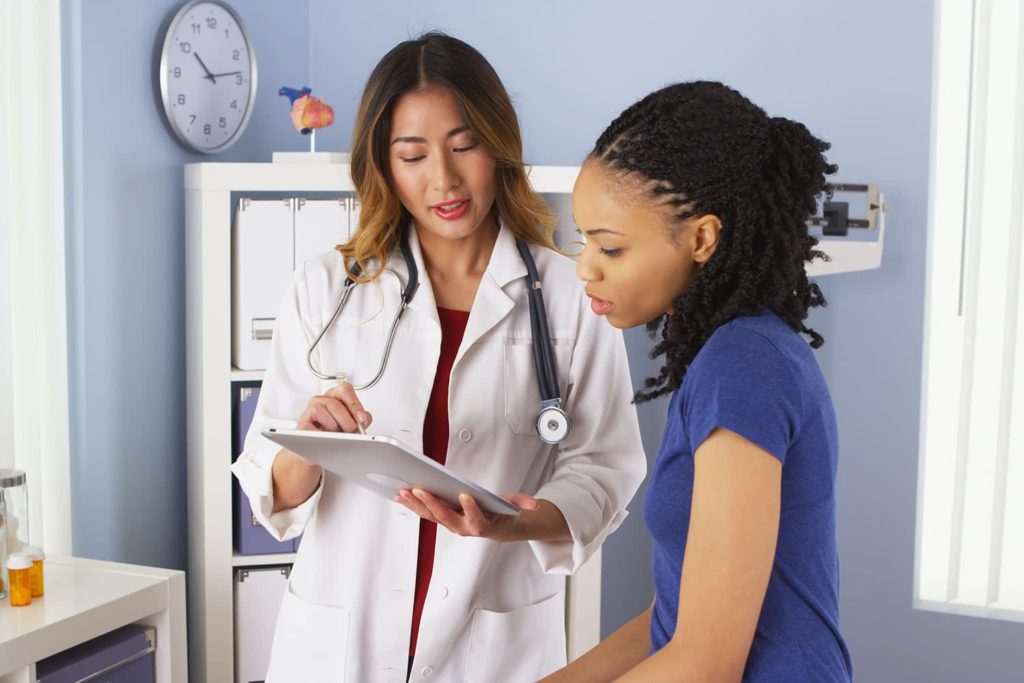 A doctor in a white coat holding a clipboard talks to a patient in a blue shirt. They are in a medical office, conveying a professional empathetic tone.