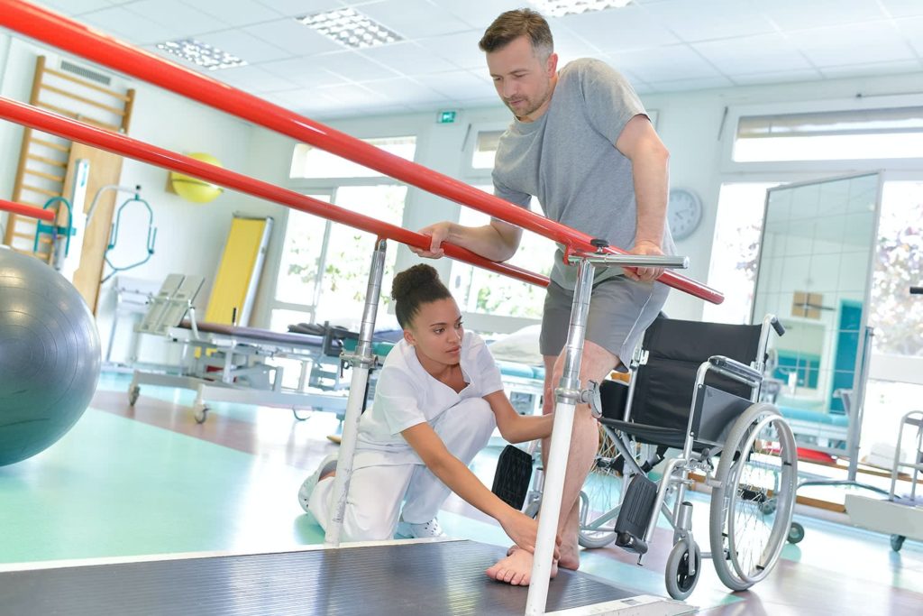 A phsiotherapist assists a man with a leg prosthetic as he practices walking between parallel bars in a rehab center, conveying determination.