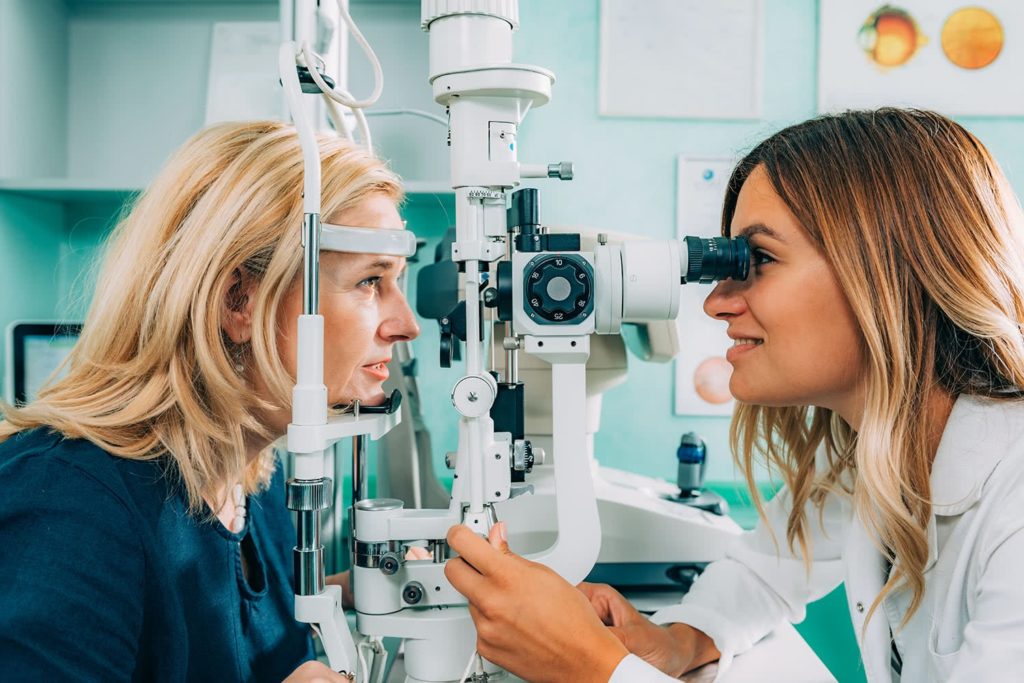 Eye exam scene with a female optometrist using a slit lamp machine to examine a patient's eyes in a clinical setting. Both appear focused and calm.