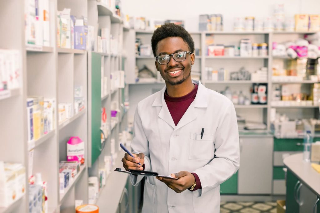 A pharmacist in a white coat smiles while holding a clipboard in a well-stocked pharmacy. The shelves behind him are filled with various medication boxes.