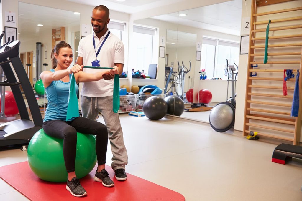 A woman sits on a green exercise ball, using a resistance band with a trainer's guidance in a bright gym. Treadmills and exercise balls are visible.