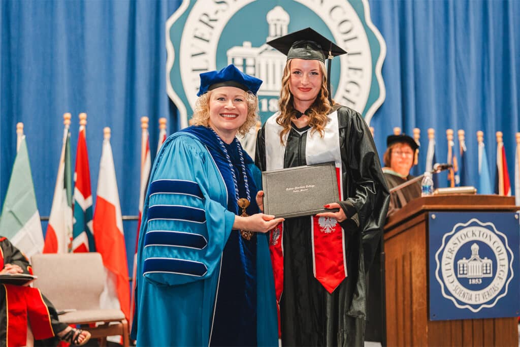 A graduate in cap and gown receives a diploma from a woman in blue academic regalia on stage. Background features flags and a college emblem.