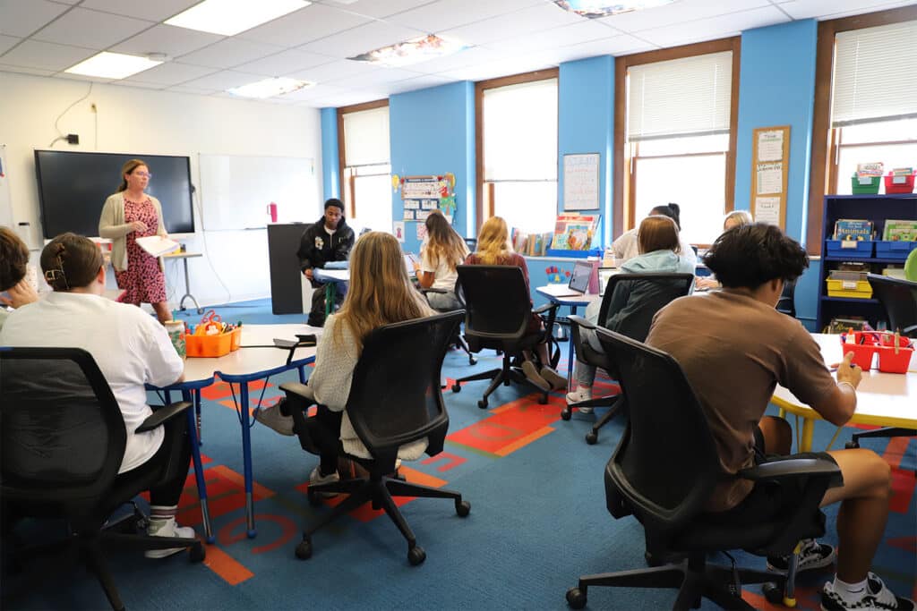 A classroom with students seated at desks in a bright, colorful room. A teacher stands at the front, engaging with the class. The atmosphere is focused and lively.