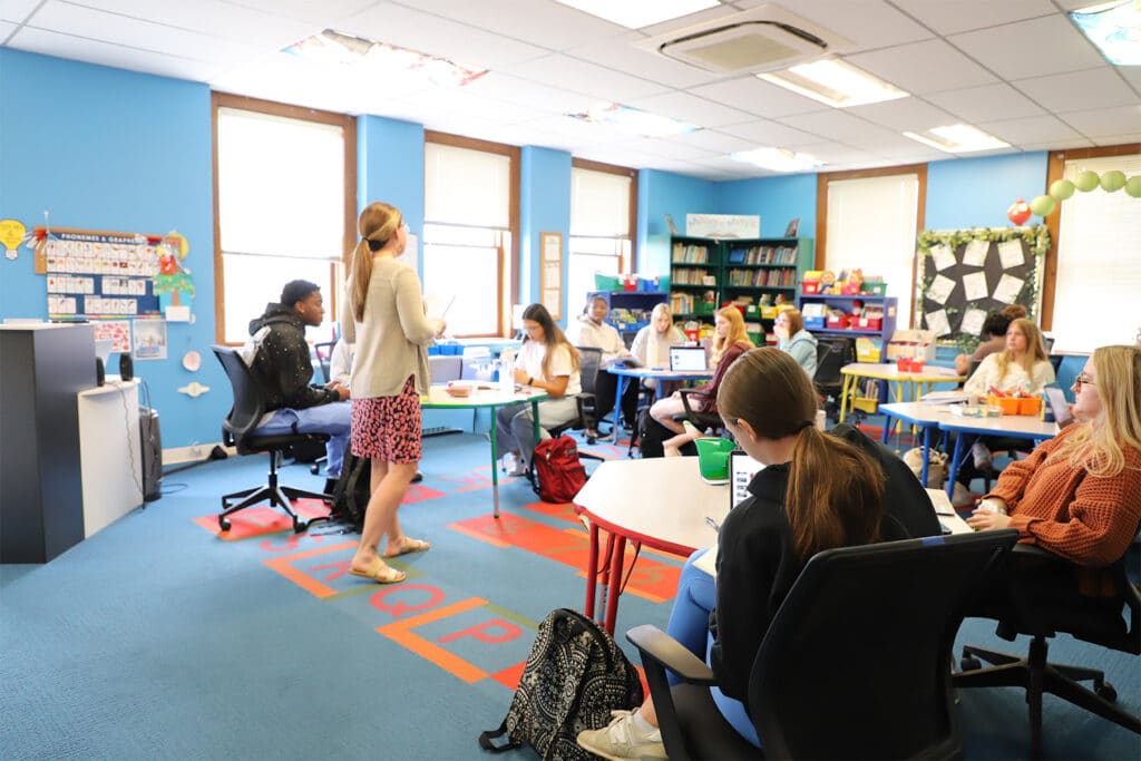 A bright lassroom with blue walls, students seated at desks, and a teacher standing, gesturing towards a whiteboard. The atmosphere is focused and engaging.