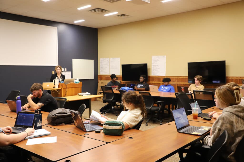 A classroom with a teacher at a podium and students seated around tables, working on laptops. The atmosphere is focused and engaged.