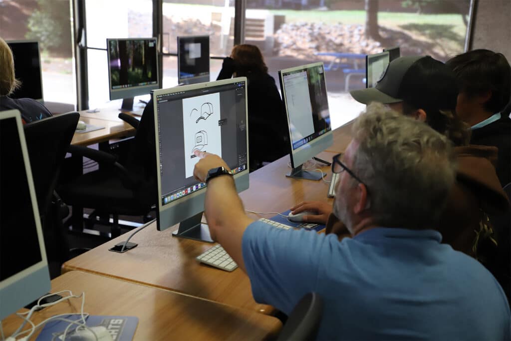 A classroom setting with a group of students at desks using desktop computers. An instructor in a blue shirt points at a screen displaying simple drawings of hats, indicating a design or art lesson. The room is bright with natural light from large windows, creating a collaborative and focused atmosphere.