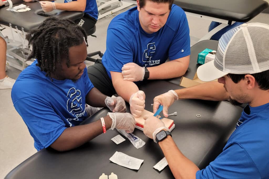 Three individuals in blue shirts practice a medical procedure on a synthetic arm. They wear gloves and appear focused and collaborative.