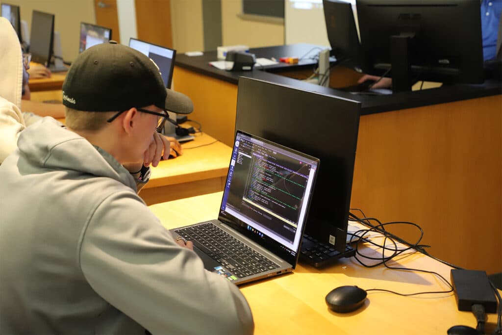 A student wearing a cap and hoodie is focused on a laptop displaying code, sitting in a classroom with several computers. The setting is educational and tech-oriented.